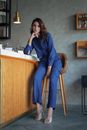 Stylish woman in blue jumpsuit poses at a sleek café with modern decor, seated elegantly on a bar stool.