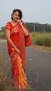 An adult woman stands on a rural road, wearing a vibrant red saree, embodying traditional elegance.