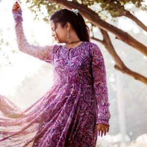 A woman poses gracefully in a vibrant purple dress, basking in sunlight amidst the trees.