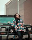 Young woman in plaid shirt leans on an off-road vehicle in an urban setting in Iran.