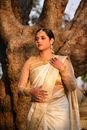 A woman in a golden saree stands gracefully by a tree, exuding cultural elegance.