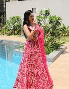 An elegant woman in a vibrant pink lehenga poses gracefully by a poolside during a sunny day.