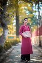Full body of feminine young Asian lady in elegant dress with ornamental fan in hand standing on path in autumn park and looking at camera